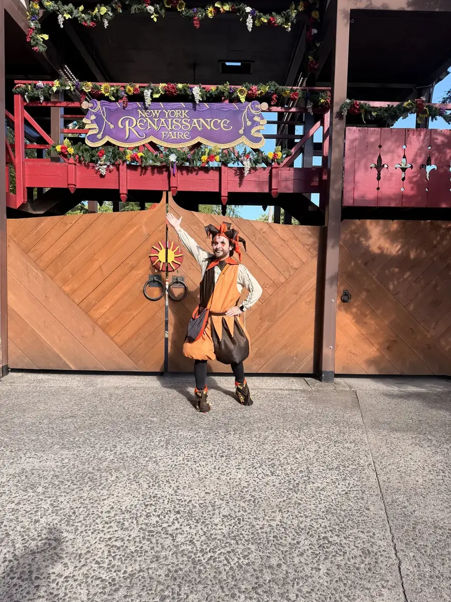 Grahm dressed as a jester in an orange and brown costume with a jester's hat, standing with one arm raised in front of the wooden gates of the New York Renaissance Faire