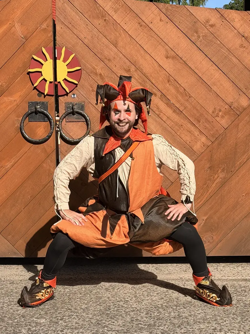 A closer view of the same jester costume, showing the orange and brown panelled tunic, the laced sleeves, the hat with bells, and the decorated shoe covers